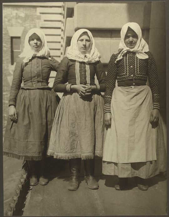 A black and white photograph of three women in traditional clothing standing in front of a stone wall.