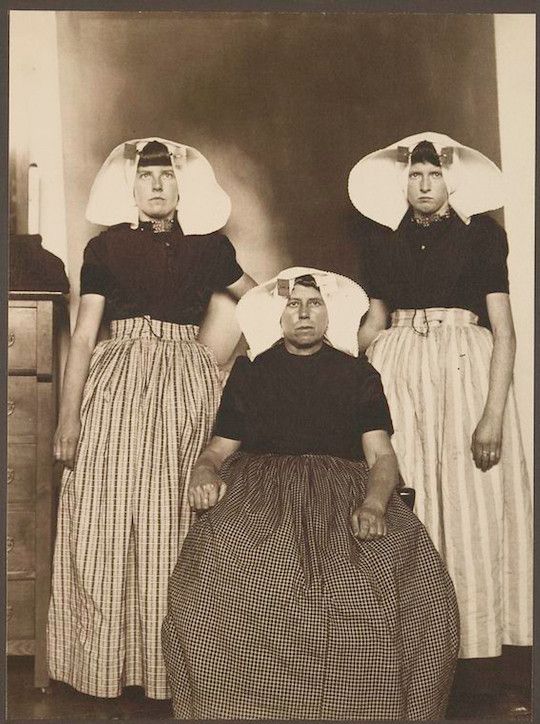 A sepia-toned photograph of three women in traditional clothing, posing in front of a simple wall.