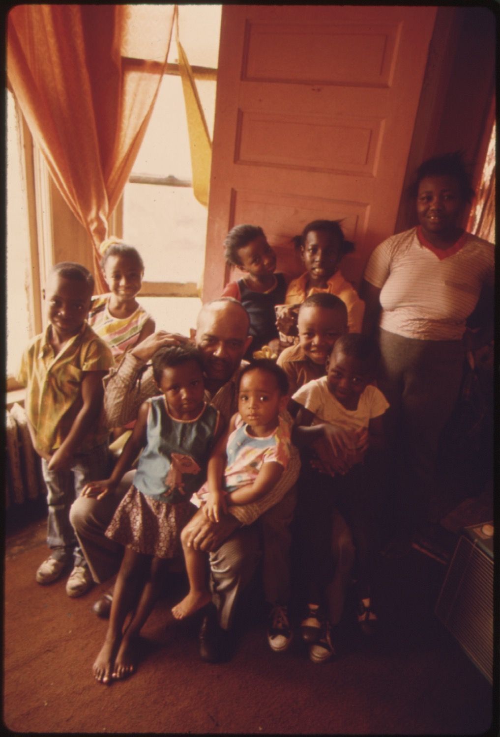 A group of children and a woman are gathered in a room with red curtains and peeling paint.