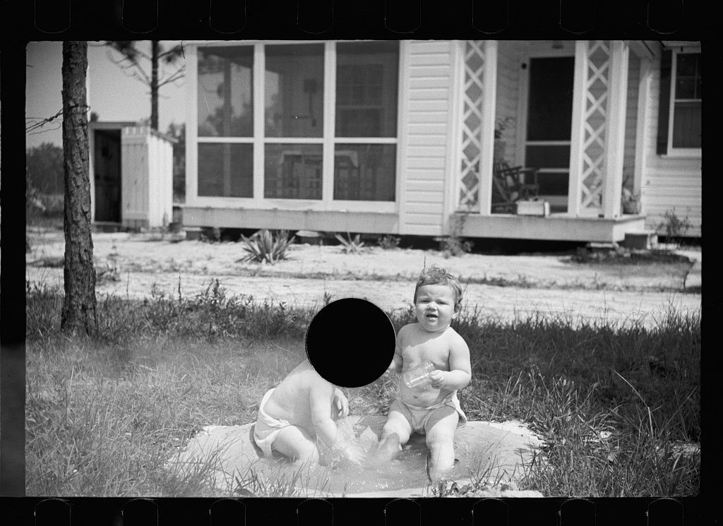 Black and white photograph of two children playing in a shallow pool of water in front of a house.