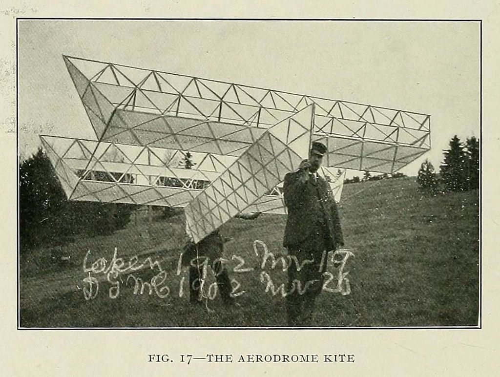 A vintage black and white photograph of a large, geometric kite being held by a person in a field.