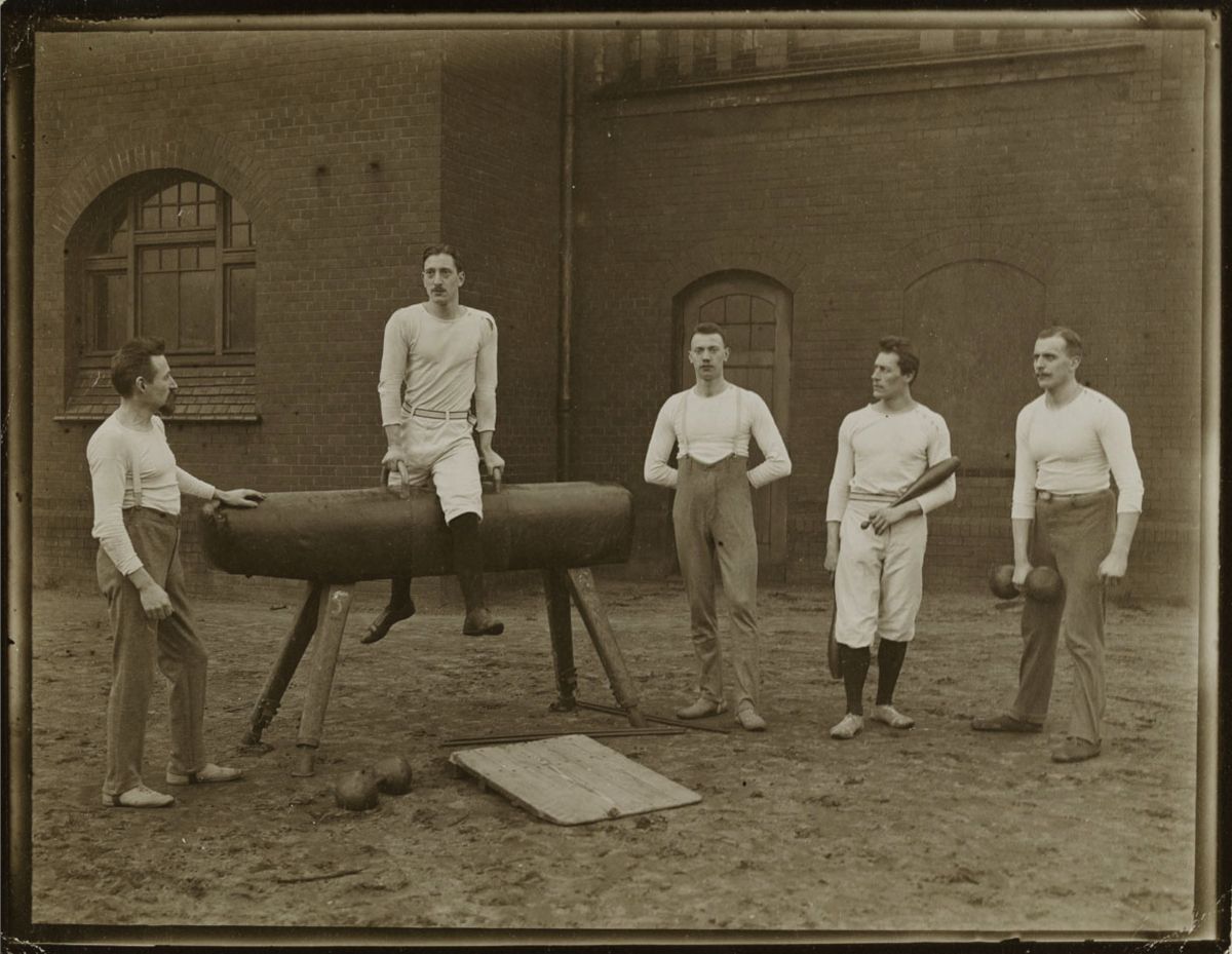 A vintage sepia-toned photograph of six men in a gymnasium or outdoor training area. One man is seated on a wooden buck, while the others stand around him holding balls or weights.