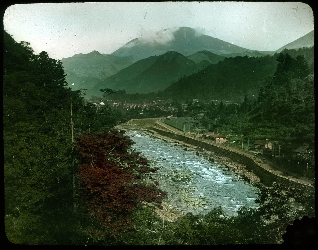 A vintage photograph of a town nestled in a mountainous landscape, with a winding river flowing through the center.