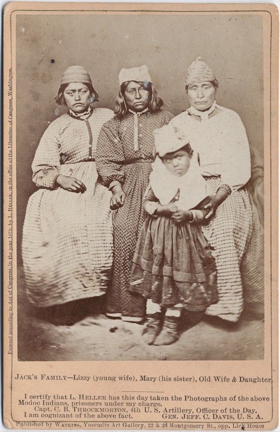 A vintage photograph of four Native American women identified as Jack's family: Lizzy (young wife), Mary (his sister), Old Wife, and Daughter. They are dressed in long patterned dresses with white aprons and head coverings. The photograph is accompanied by text certifying that the subjects are Modoc Indians who were prisoners under the charge of Captain C. B. Throckmorton and General Jeff. C. Davis.