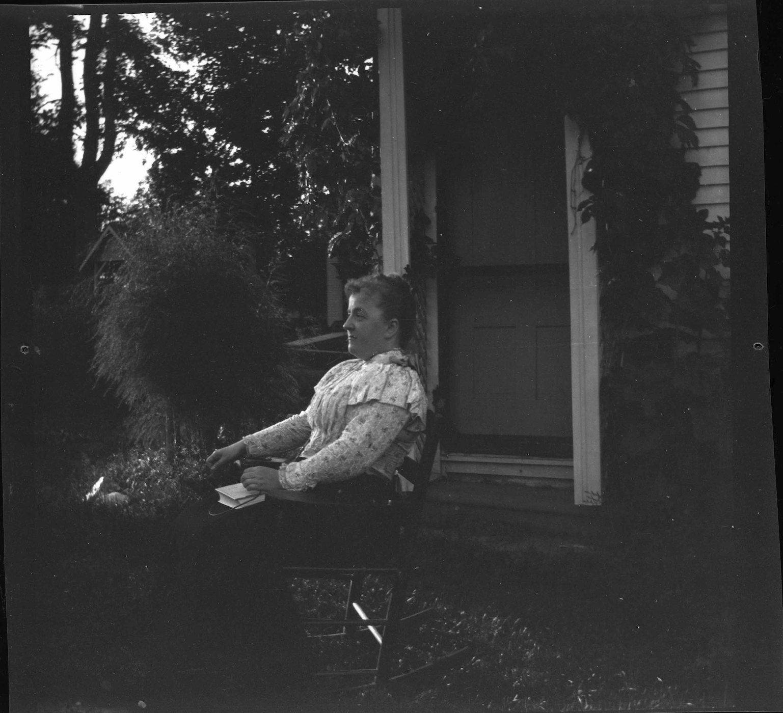 A black and white photograph of a woman sitting in a chair, reading a book next to a house.