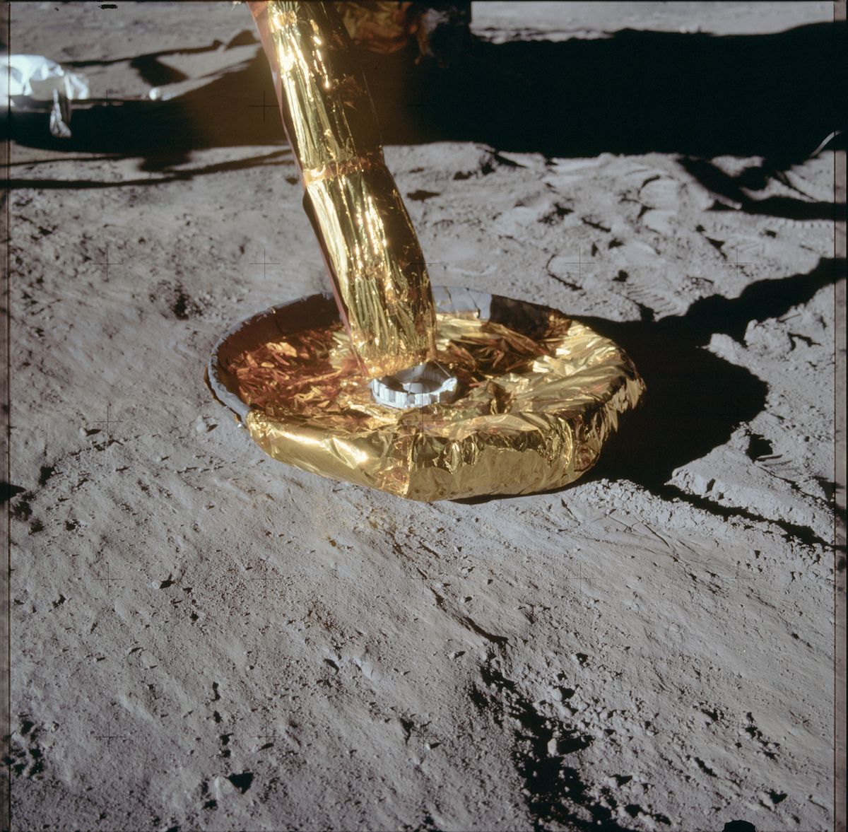 Close-up of an astronaut's boot on the lunar surface, covered in golden reflective material, with a metallic ring at the base and a strong shadow cast by the sun.