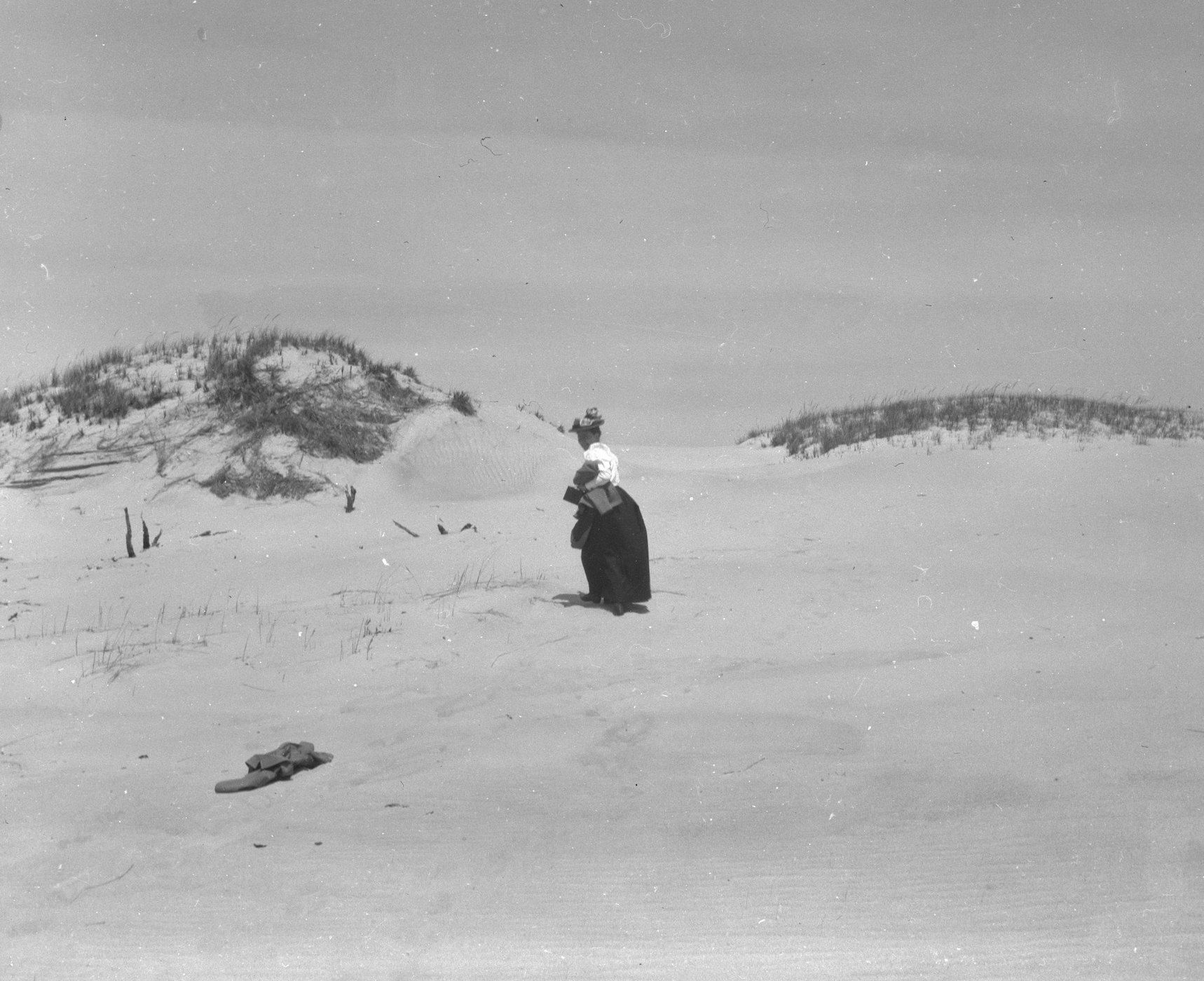 A black and white photograph of a person standing on a sandy beach, with sand dunes in the background and a pair of shoes in the foreground.