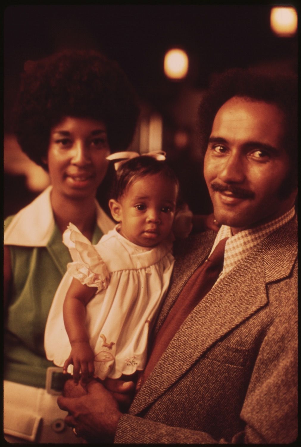 A vintage-style photograph of a man holding a baby while a woman stands behind them. The image has a warm, sepia-toned aesthetic.
