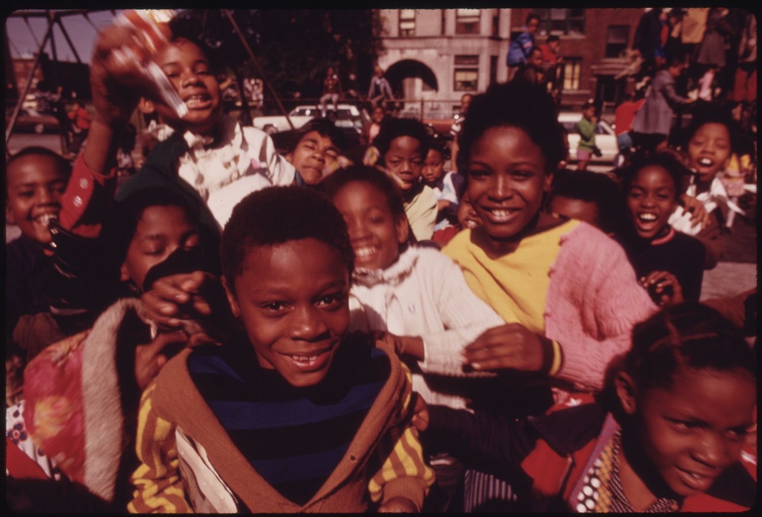 A close-up photograph of a large group of African American children smiling and gathered on a street. The children are densely packed, their faces beaming with joy and excitement. A young boy in the foreground is prominently featured, looking directly at the camera with a bright smile. The background is filled with other children, some waving their arms, creating a sense of movement and liveliness. The setting appears to be an urban street, with buildings and cars visible in the background. The image has a slightly grainy texture, which adds to its vintage feel.