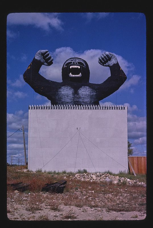 A large, black gorilla is painted on a white wall, with its arms raised and muscles flexed. The wall is topped with a row of triangular spikes, and the background is a bright blue sky with scattered clouds.