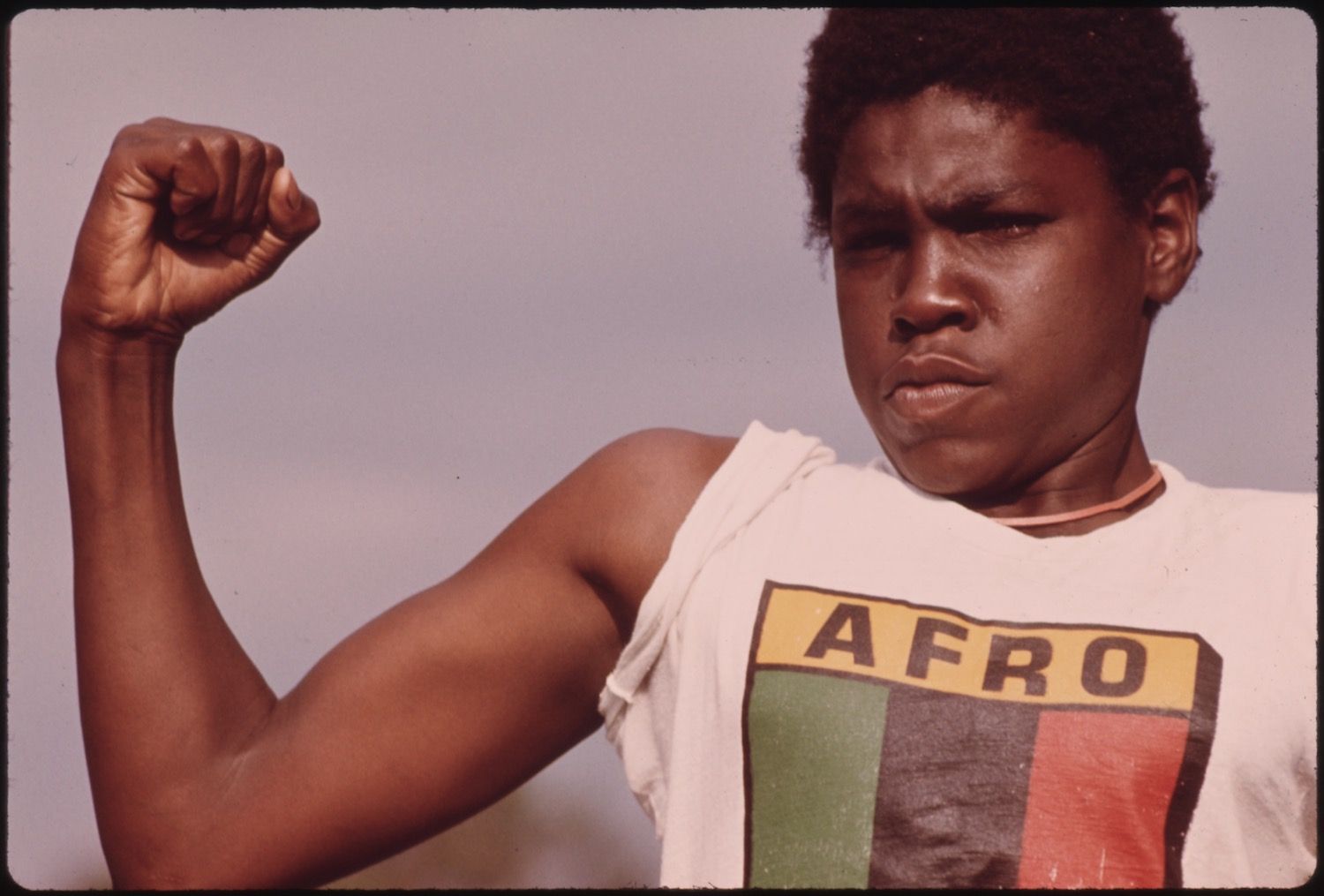 Close-up portrait of a Black man flexing his arm with a clenched fist, wearing a white tank top with the word 'AFRO' and Pan-African colors.