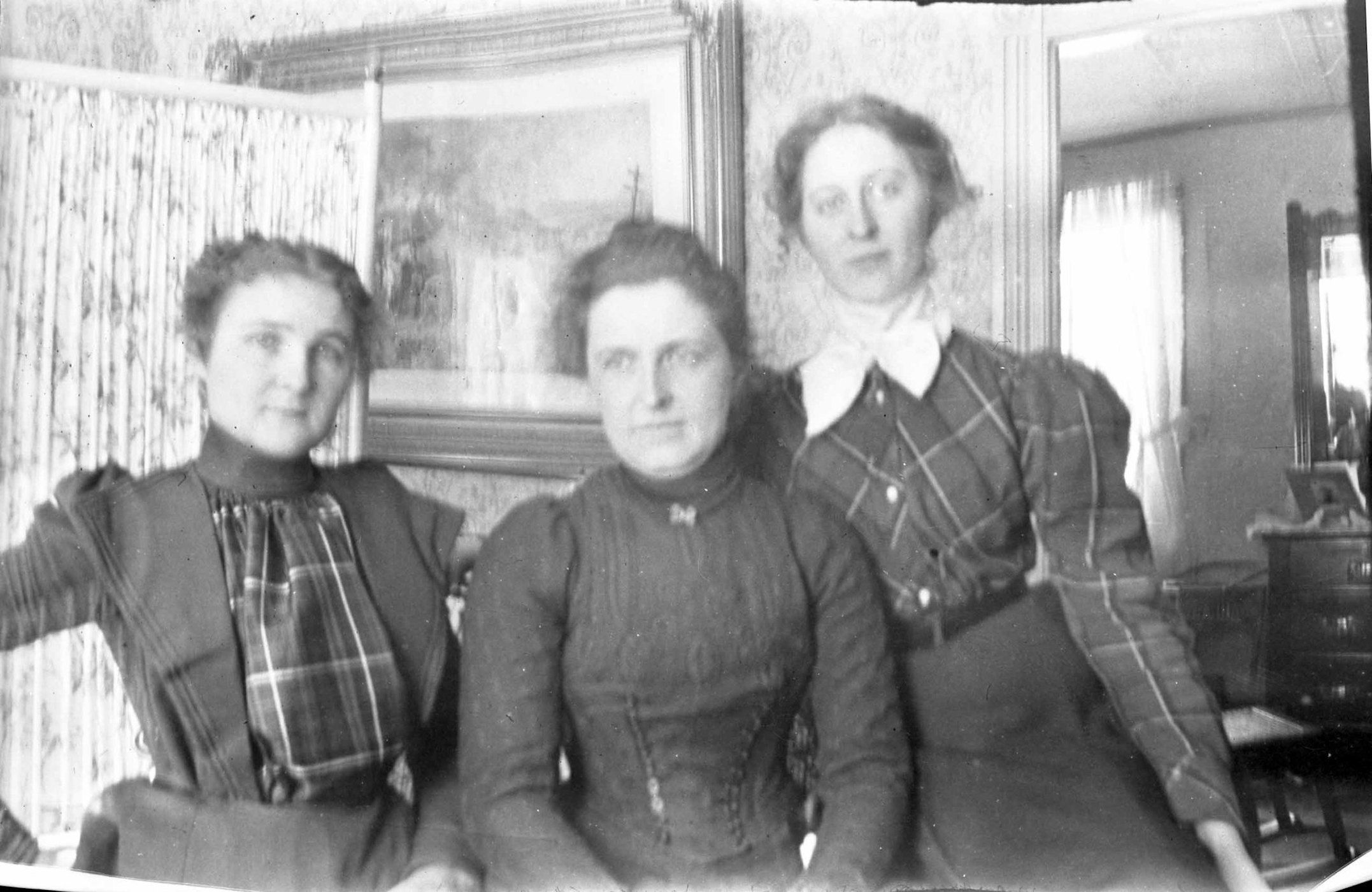 A vintage black and white photograph of three women in period clothing posing indoors.