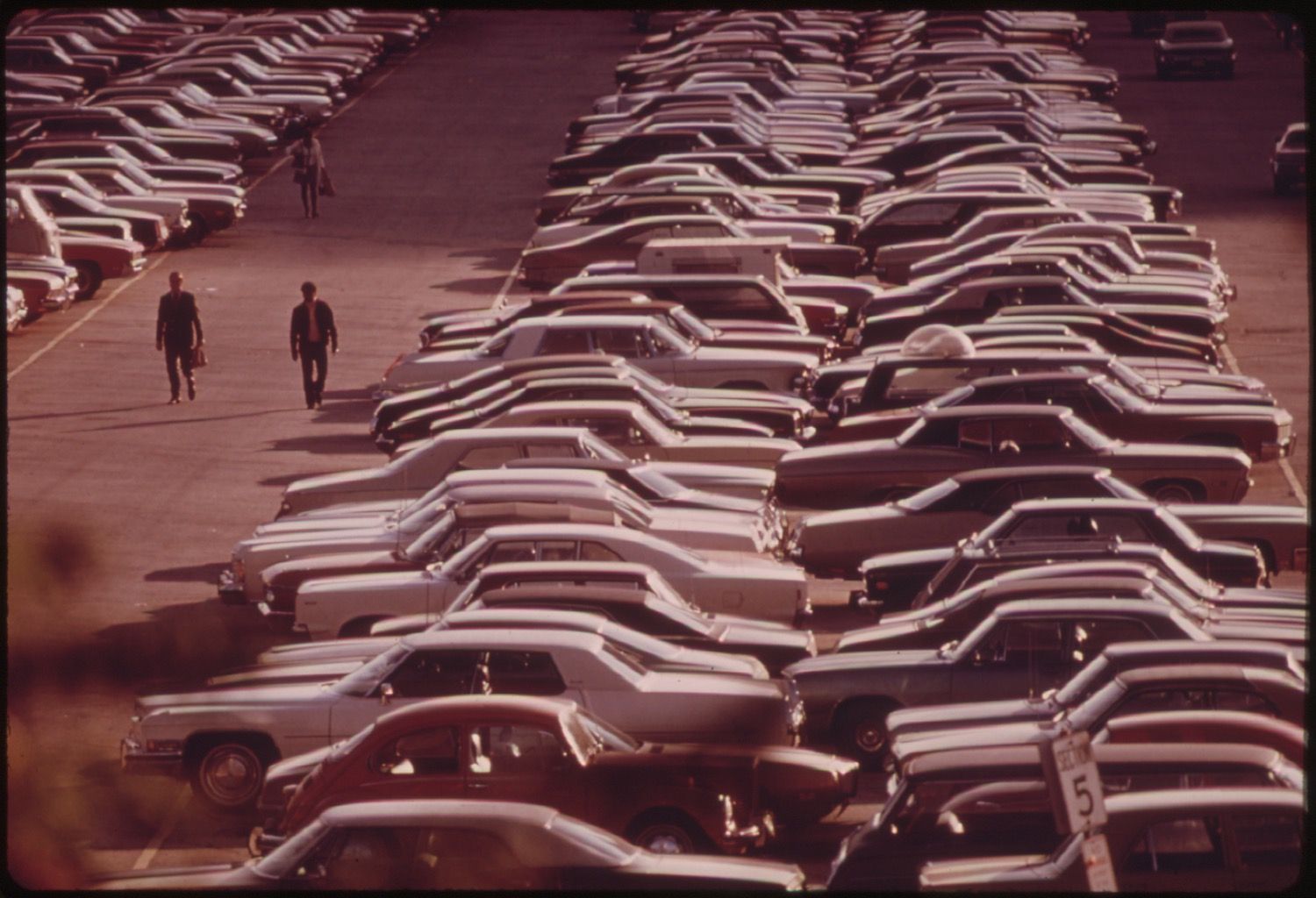 A high-angle view of a parking lot filled with rows of identical cars, with two men walking between them.