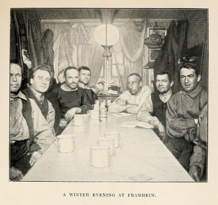 A black and white illustration of eight men seated around a long table in a rustic room, titled "A Winter Evening at Framheim."