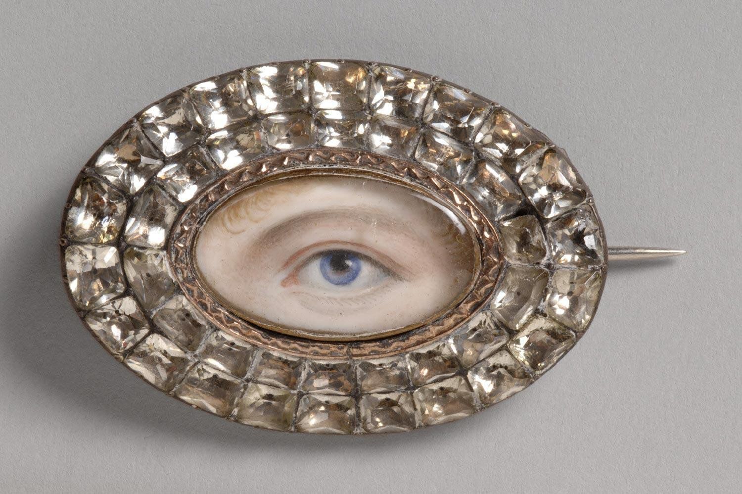 Close-up of an antique oval brooch featuring a realistic painting of a human eye surrounded by a border of clear gemstones.