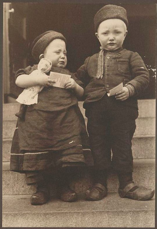 A vintage black and white photograph of two young children, one crying and holding a doll, the other stoic, both holding cards and standing on outdoor steps.