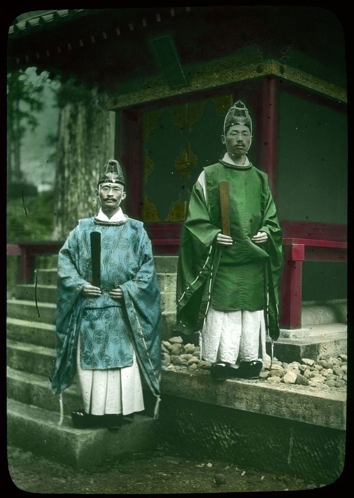 Two men in traditional Chinese attire standing on a stone staircase in front of a building with a red wooden structure.
