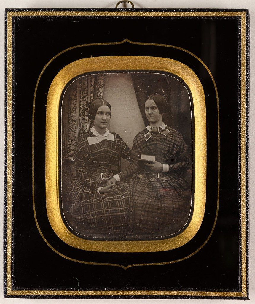 A daguerreotype portrait of two women in matching plaid dresses, seated and holding a book, encased in a decorative frame.