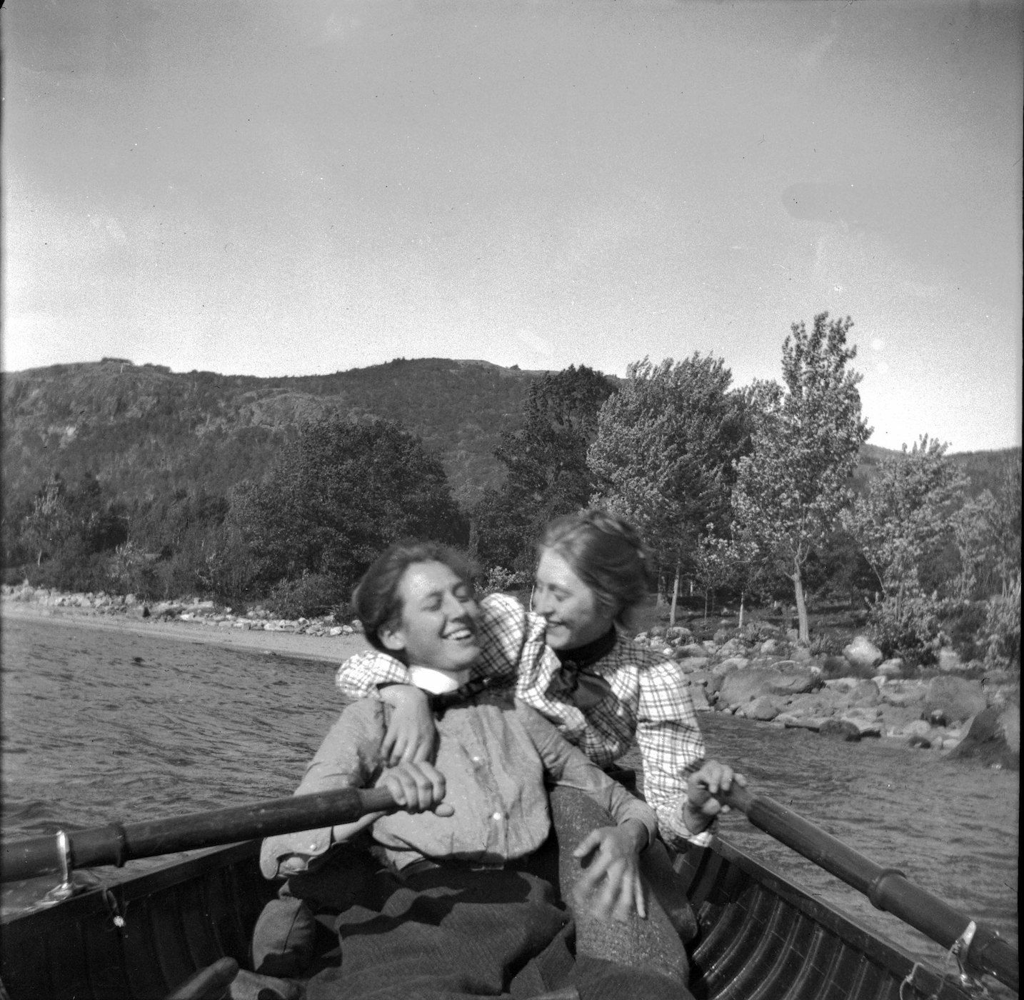 A black and white photograph of two women in a rowboat, smiling and whispering to each other. They are surrounded by trees and a mountain range.
