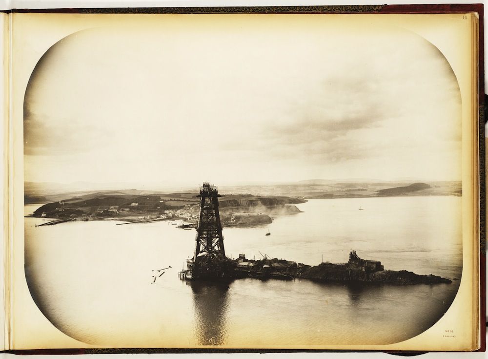 Vintage sepia-toned photograph of a coastal scene with a tall tower and complex dock structure extending into the water.