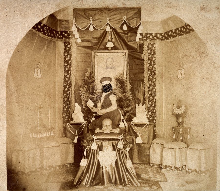 A sepia-toned vintage photograph of a skull displayed on a draped table, adorned with jewelry and a book, under a canopy-like structure with a portrait in the background.