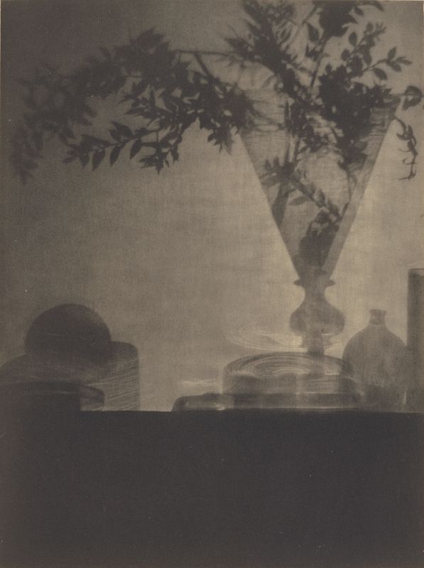 A vintage black and white photograph of a still life arrangement featuring a tall vase filled with foliage, a stack of plates, a bowl, and a bottle.