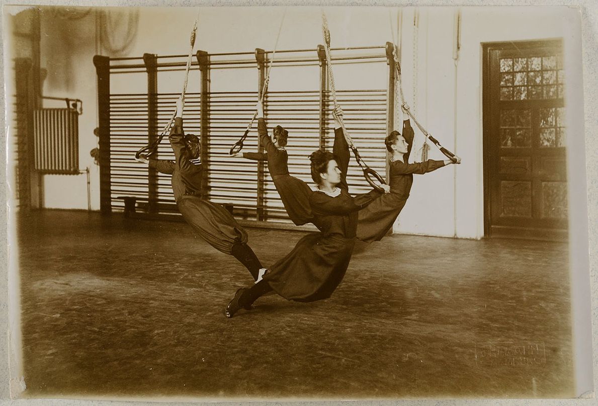 Sepia-toned photograph of three gymnasts suspended in the air with ropes in a gymnasium.