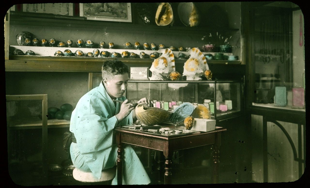 Vintage photograph of a man working at a table in a shop or studio, surrounded by shelves of decorative items.