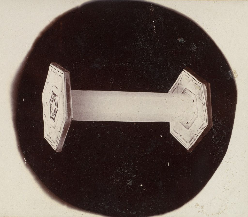 A vintage black and white photograph of a geometric object shaped like an 'H' or dumbbell, with ornate, multi-faceted ends connected by a straight bar, set against a black circular background.