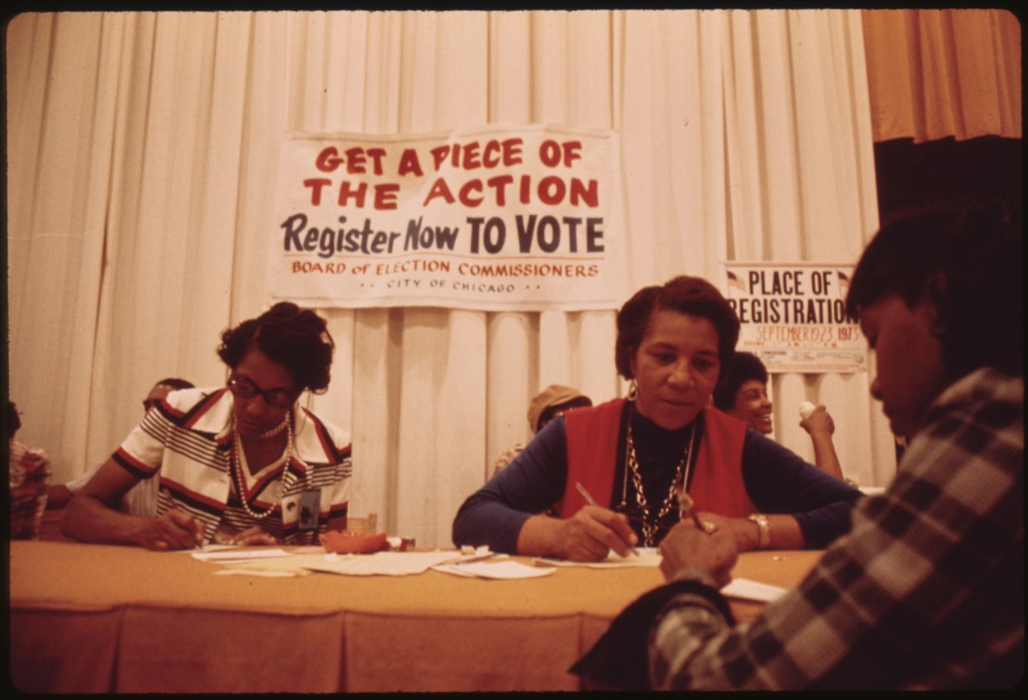 A vintage photo of African American women working at a voter registration drive in Chicago. They are seated at a table with a banner that reads "GET A PIECE OF THE ACTION Register Now TO VOTE".