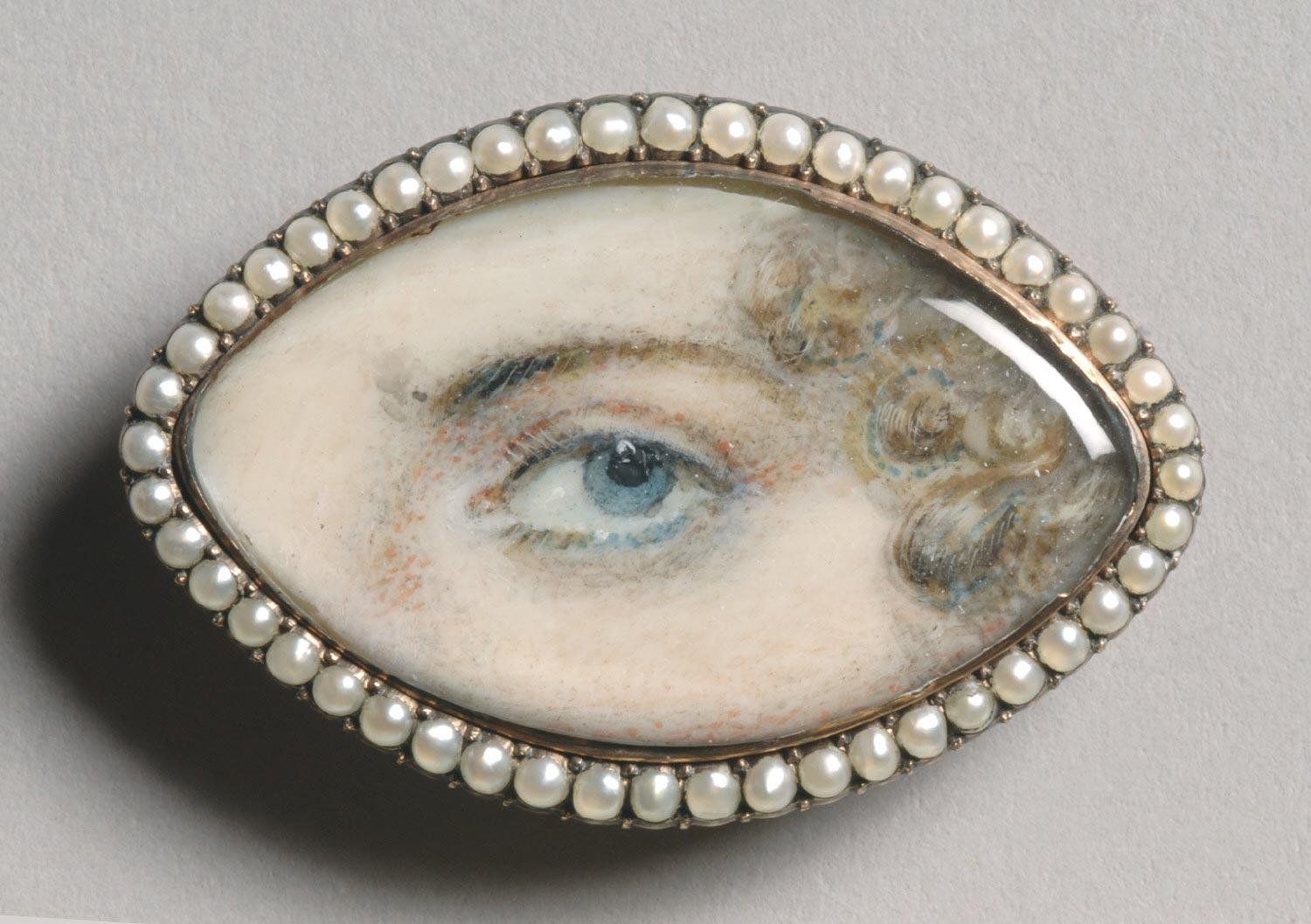 Close-up of an oval brooch featuring a miniature painting of a blue eye and curly hair, surrounded by a pearl border.