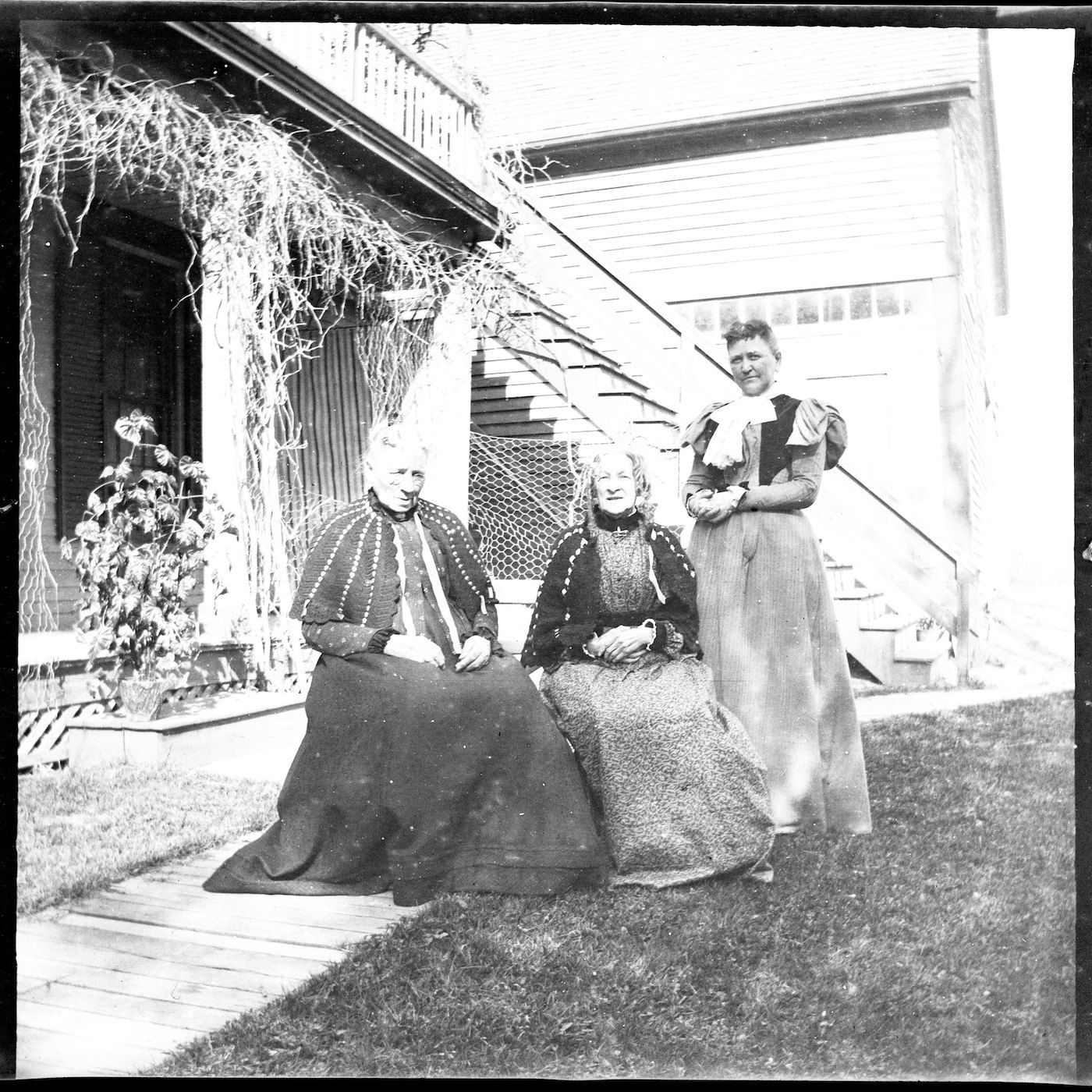 Black and white photograph of three women in long dresses posing in front of a house with stairs and a porch.