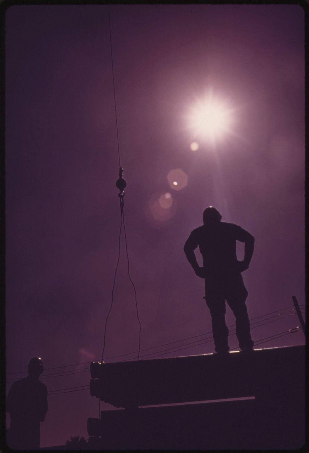 A silhouetted figure stands with hands on hips on a low wall, with another silhouetted figure to the left and a wire running vertically through the frame against a hazy, bright sky.
