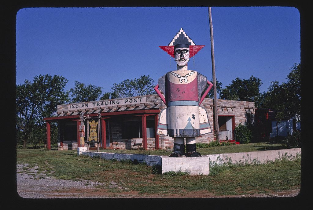 A vintage roadside attraction featuring a large, weathered statue of a Native American figure in front of an 'Indian Trading Post' building.