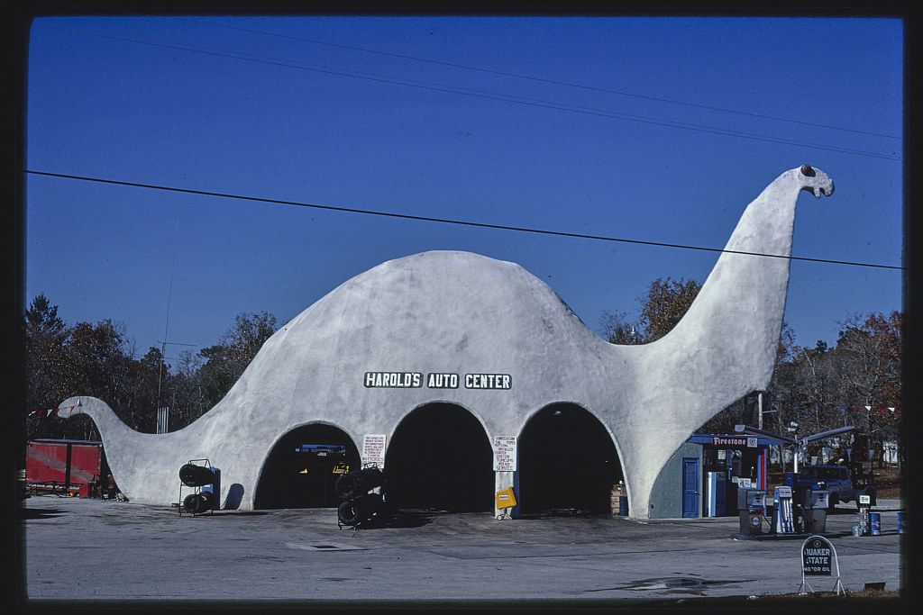 A large, dinosaur-shaped building houses Harold's Auto Center. The building is light gray and has three arched openings for bays. A paved parking lot surrounds the building, and trees and a blue sky are in the background.