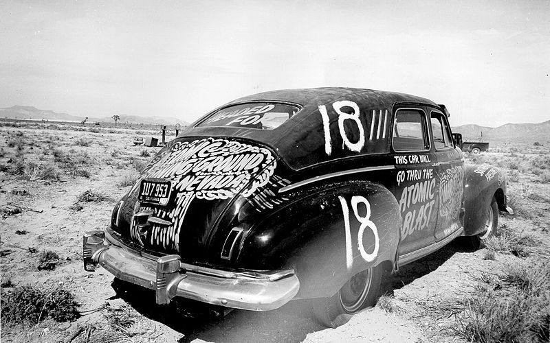 A black and white photograph of a vintage car covered in white markings and numbers in a desolate desert landscape.