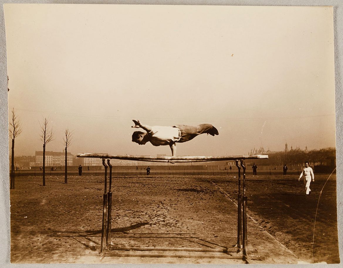 A sepia-toned vintage photograph of an athlete performing a high jump over a wooden bar in a field, with spectators and buildings in the background.