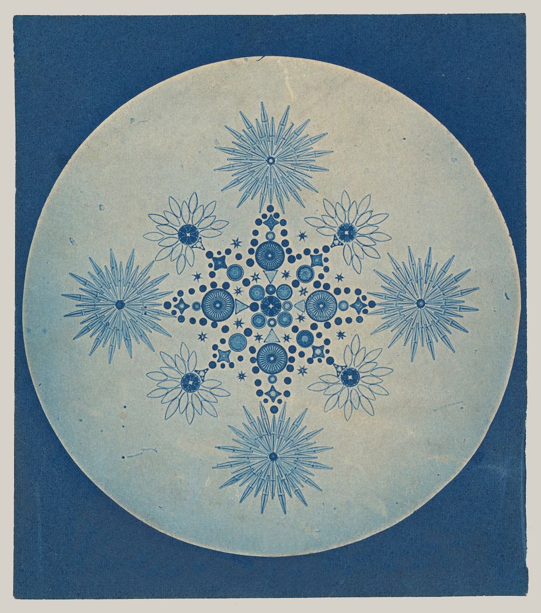 A circular design with a vintage, almost blueprint-like aesthetic. The background is a pale, muted green, resembling aged paper. Within the circle, a symmetrical pattern of floral and star motifs is rendered in a faded blue.