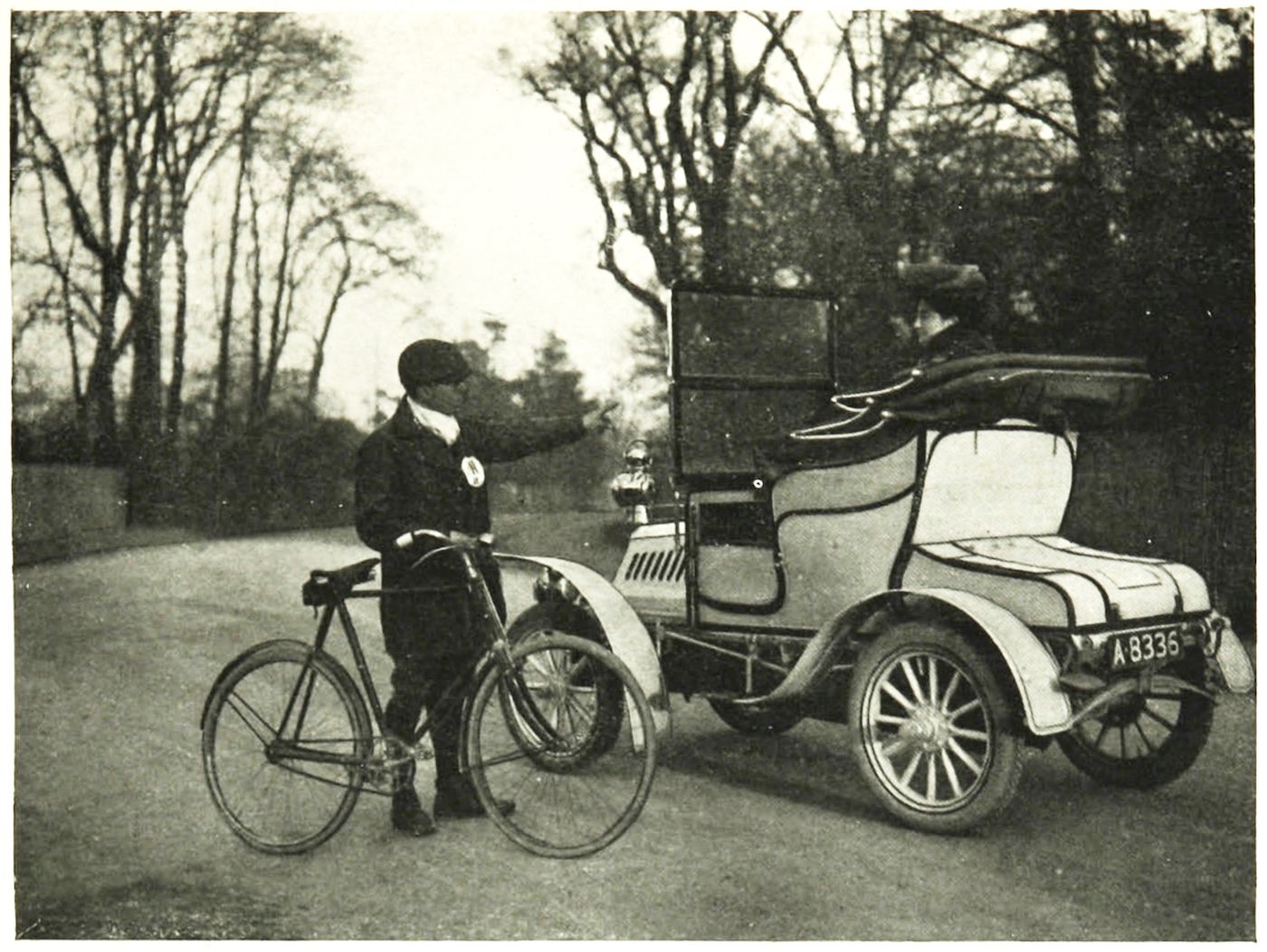 A vintage black and white photograph of a man with a bicycle next to an early model car on a dirt road, surrounded by trees.