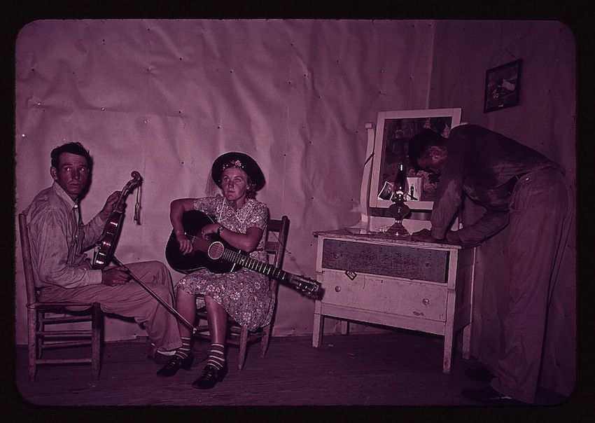 A black and white photograph with a pinkish hue, depicting a scene inside a simple room. Two people are seated on wooden chairs, playing musical instruments. The person on the left is a man, holding a fiddle and appearing to play it. The person on the right is a woman, seated and playing a guitar. To the right of the musicians is a white dresser with a mirror attached. A person, mostly obscured from view, is standing near the dresser, seemingly looking at their reflection in the mirror.