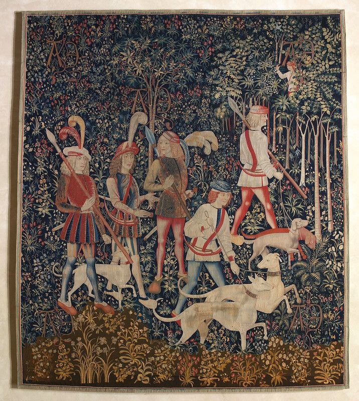 A detailed tapestry depicting a hunting scene in a dense forest with several hunters and a pack of white hunting dogs.