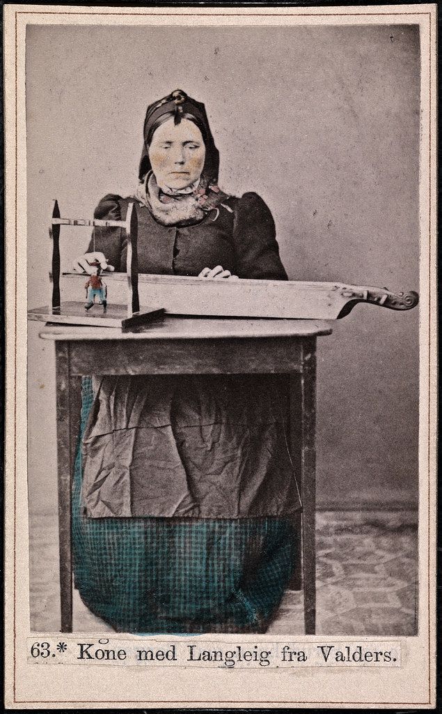 A vintage black and white photograph of a woman seated at a table with a small toy or figure on it.