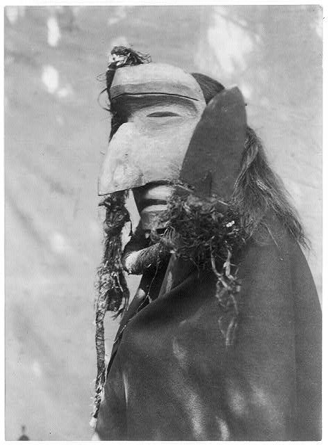 A black and white photograph of a person wearing a mask and feathered headgear.