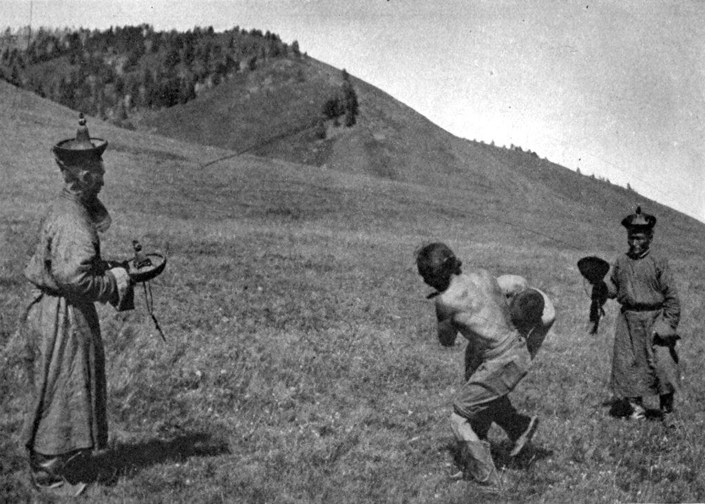 A black and white photograph of a man running with his hands tied behind his back, pursued by two figures in traditional clothing on a grassy hillside.