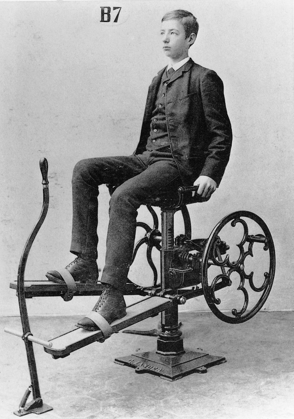 Vintage black and white photograph of a young boy on an early exercise machine.
