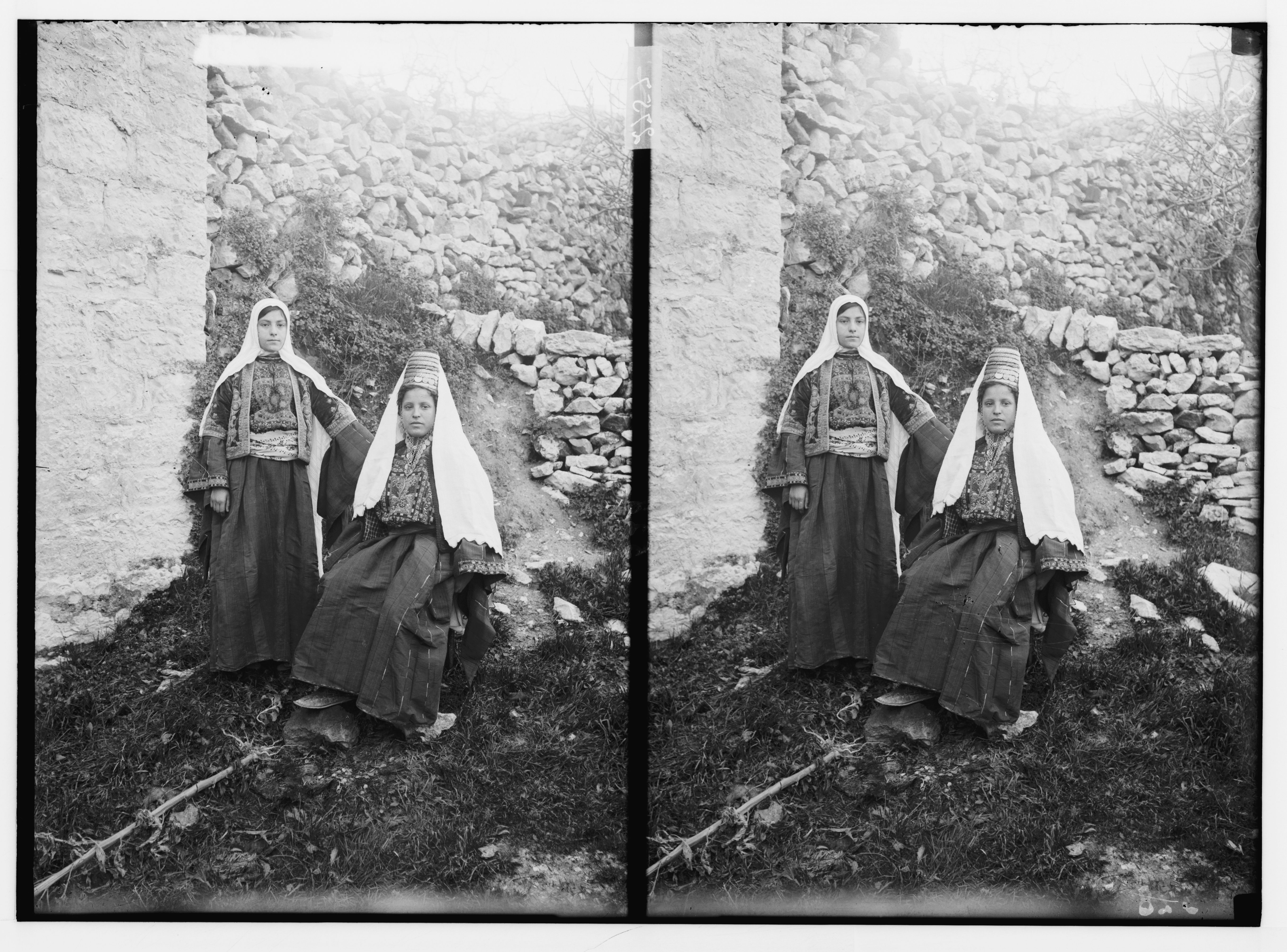 Stereoscopic photograph of two women in traditional clothing standing in front of a stone wall.