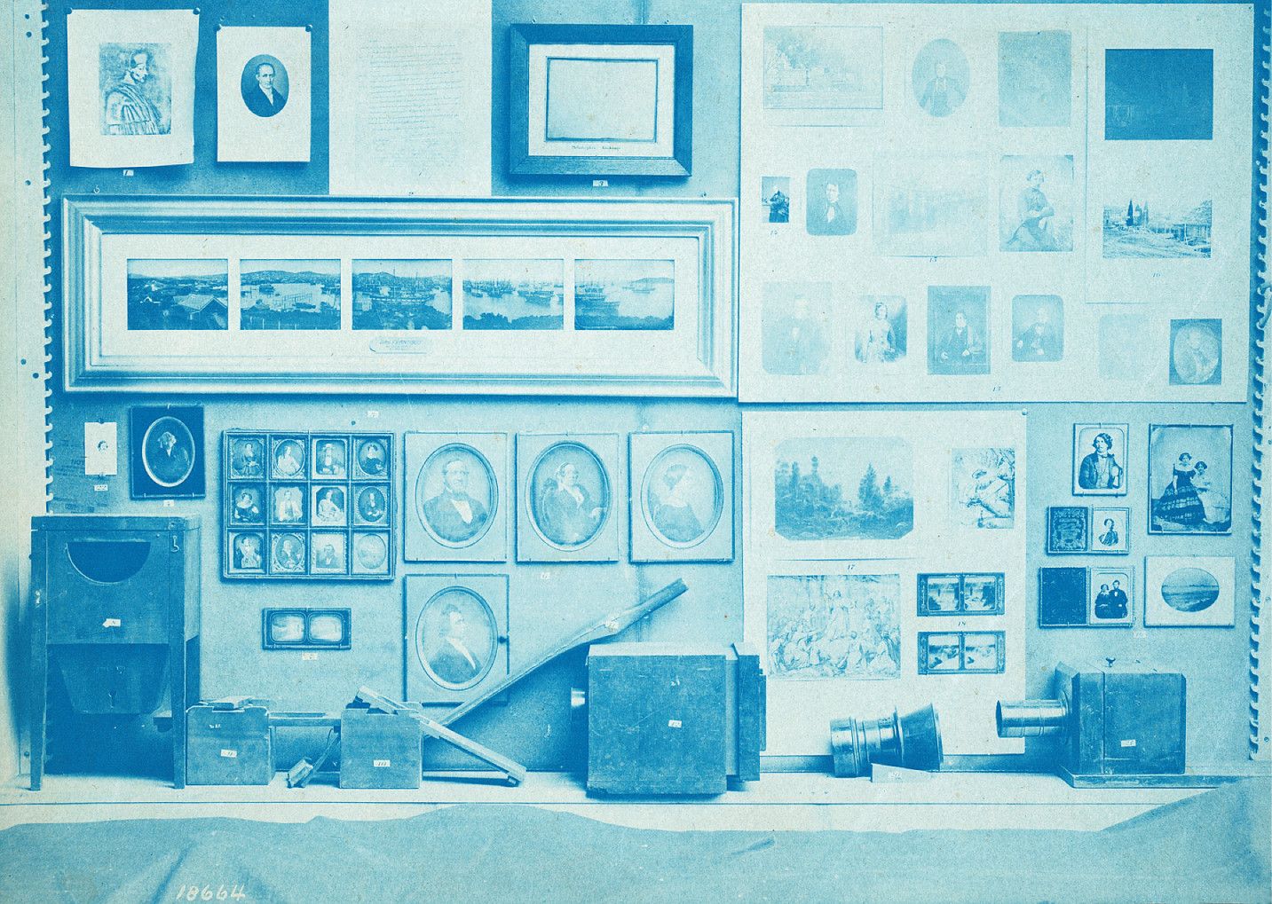 A vintage cyanotype photograph of a photographic studio interior, with walls covered in framed paintings and photographs, and the floor cluttered with trunks, boxes, and photographic equipment.