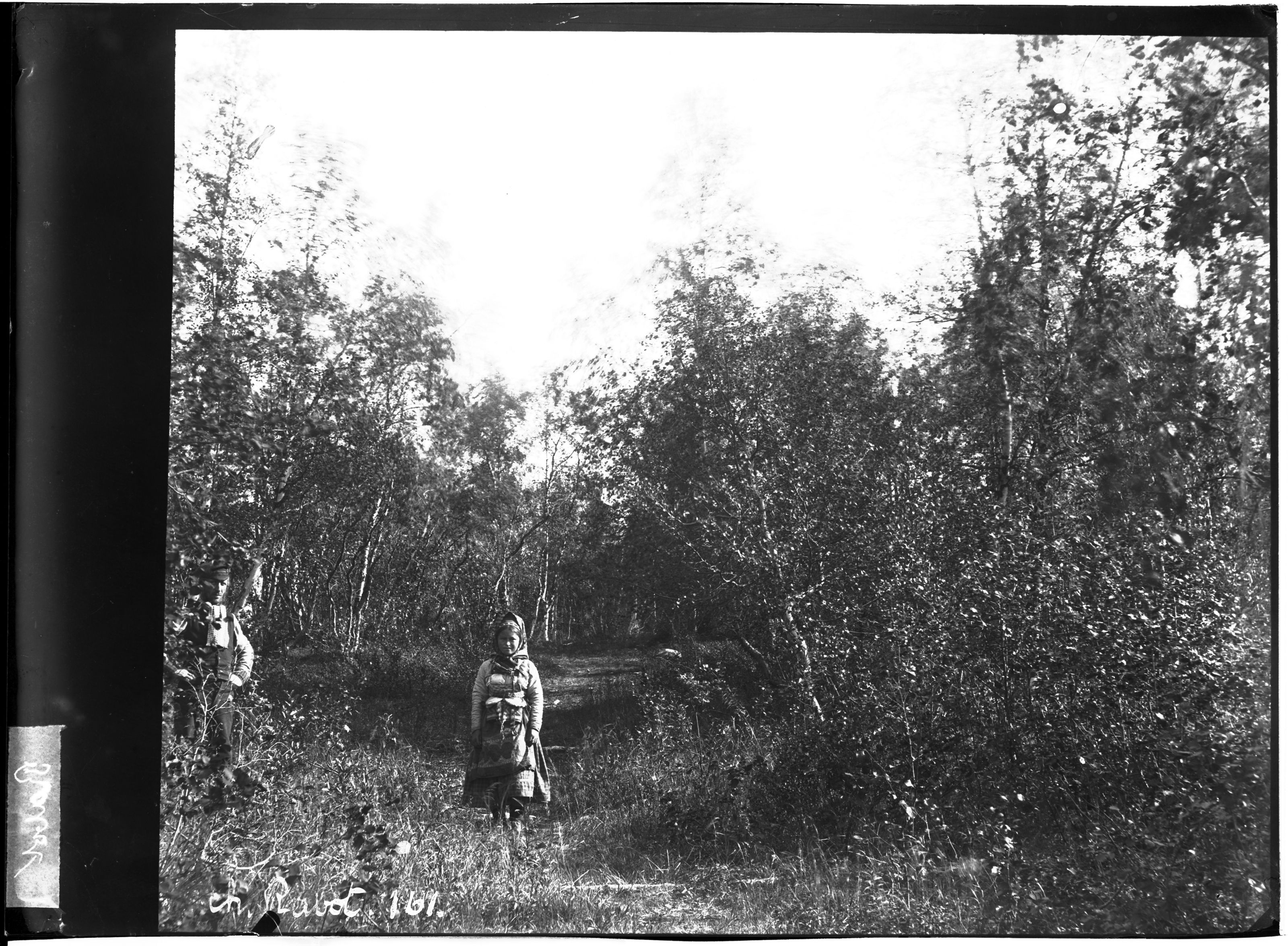 A black and white photograph of two people standing in a wooded area, with a narrow path winding through the trees.