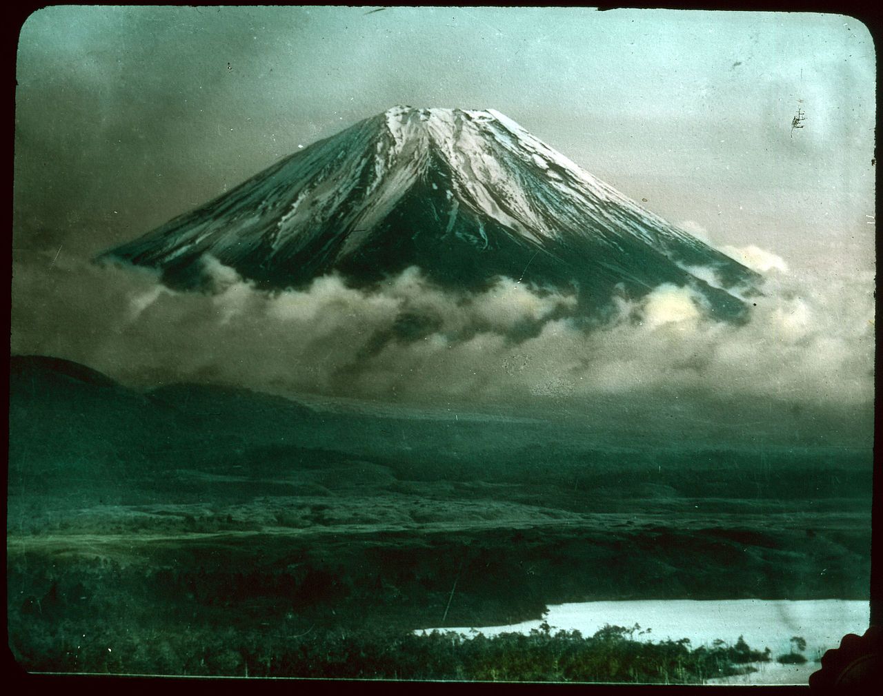 A vintage photograph of Mount Fuji, a snow-capped volcano, rising above a landscape of rolling hills and valleys.