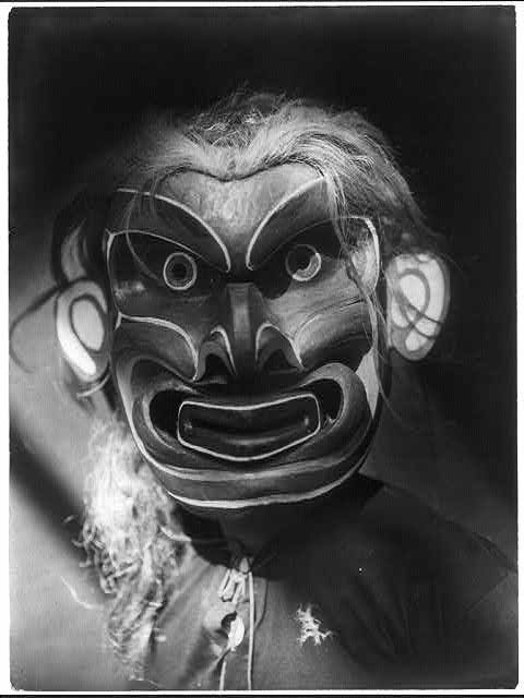A black and white photograph of a person wearing a traditional mask with exaggerated features.